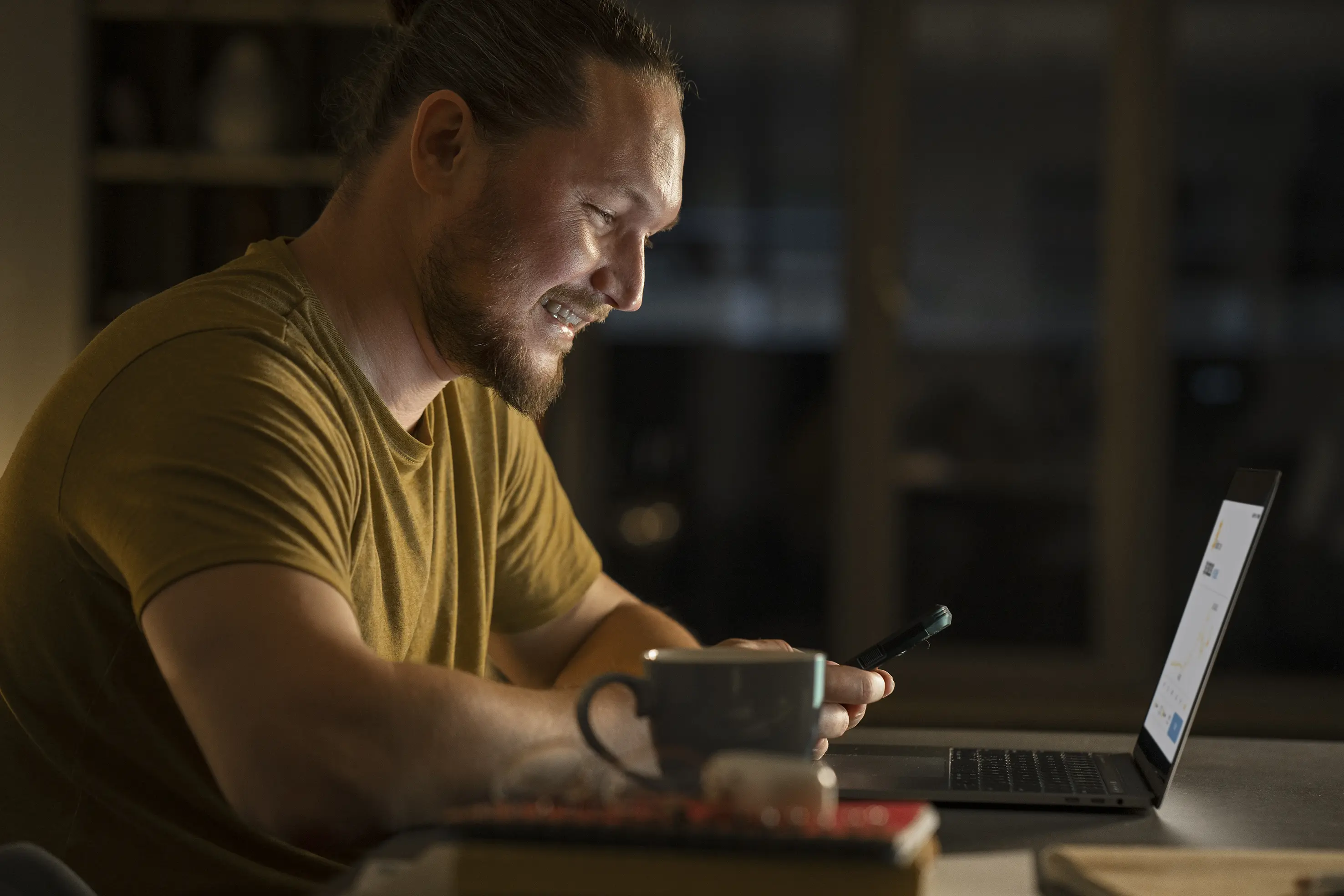 Persoon die lacht terwijl hij op een laptop werkt en een gsm in de hand heeft